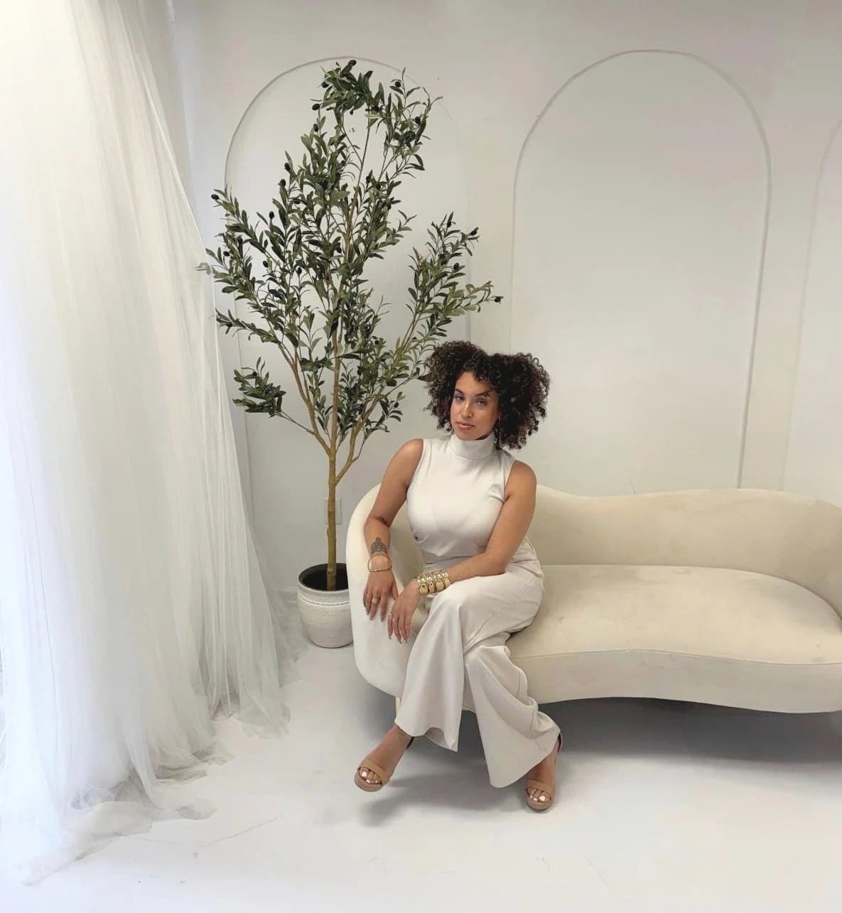 Jhay, Founder & Creative Director of You're The Alchemist, seated on a cream sofa in a bright white studio beside an olive tree